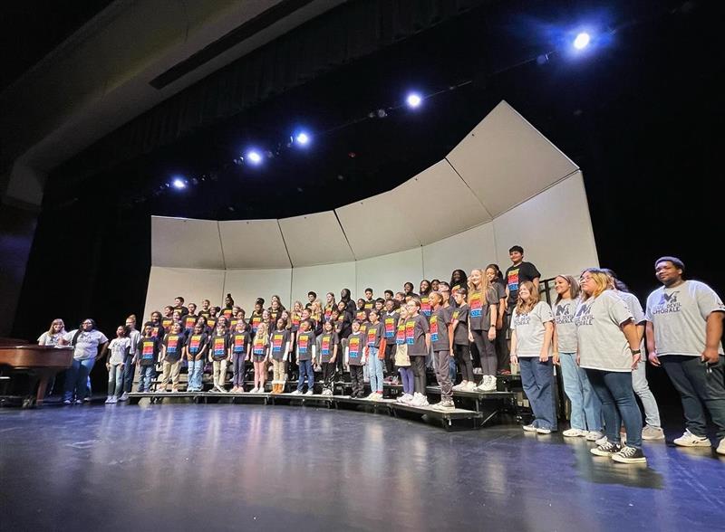 A group of young students stand on chorus risers on stage while singing